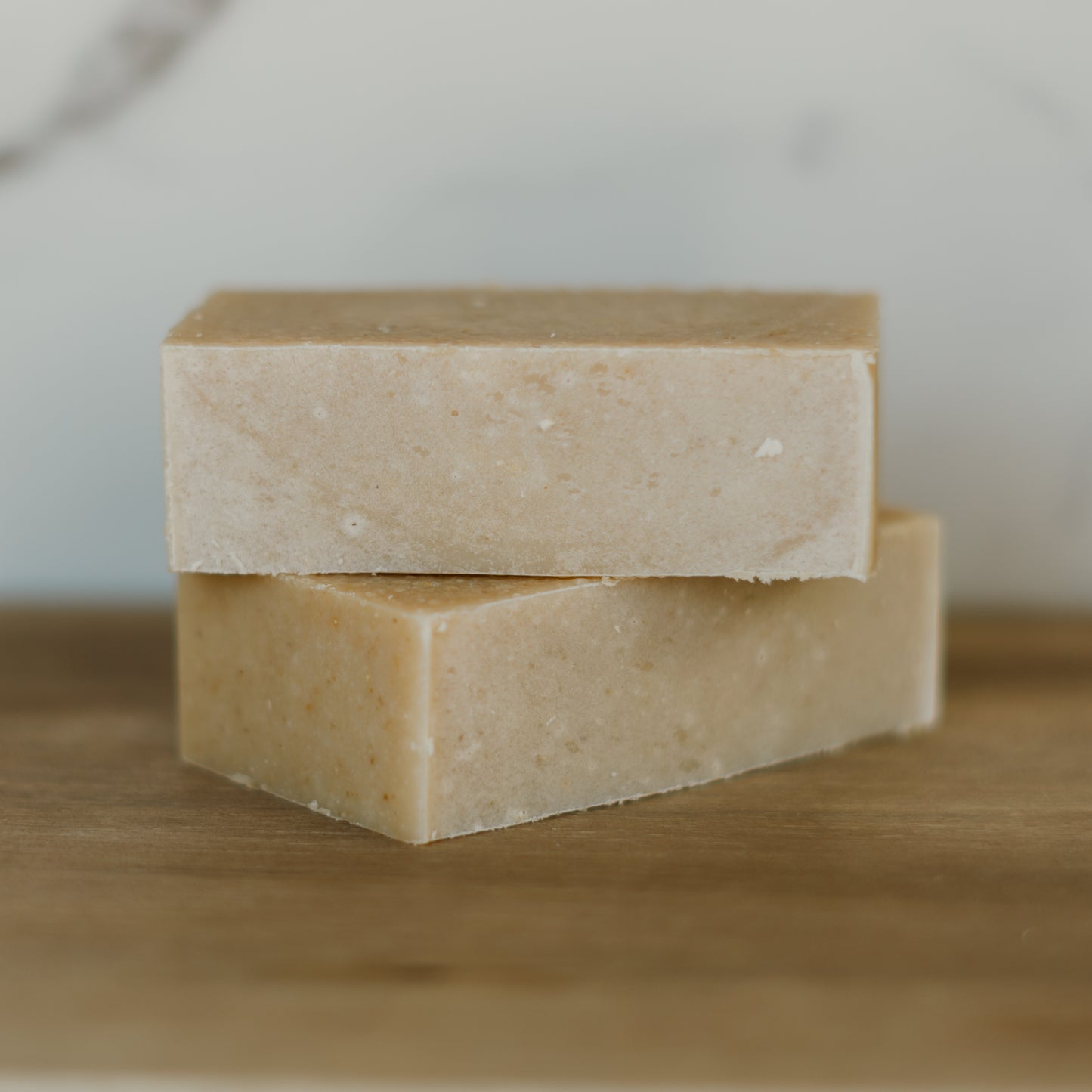 Two bars of beige soap stacked on a wooden surface with a blurred background